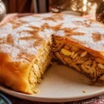 A golden Moroccan chicken bastilla with a sliced wedge, powdered sugar, cinnamon, and almonds.