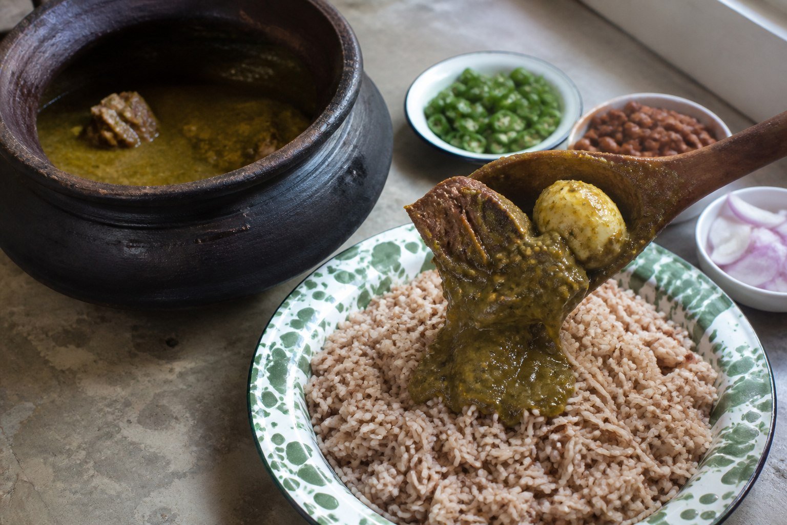 Ayamase stew with beef and egg being spooned over loose brown Ofada rice from a clay pot.