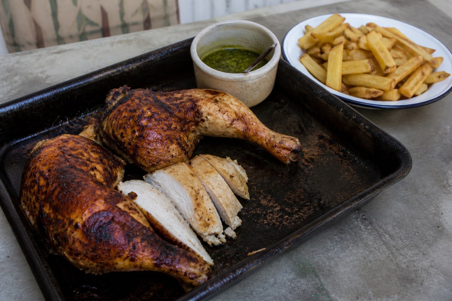 Roasted Peruvian chicken being carved on a tray beside fries and green sauce.
