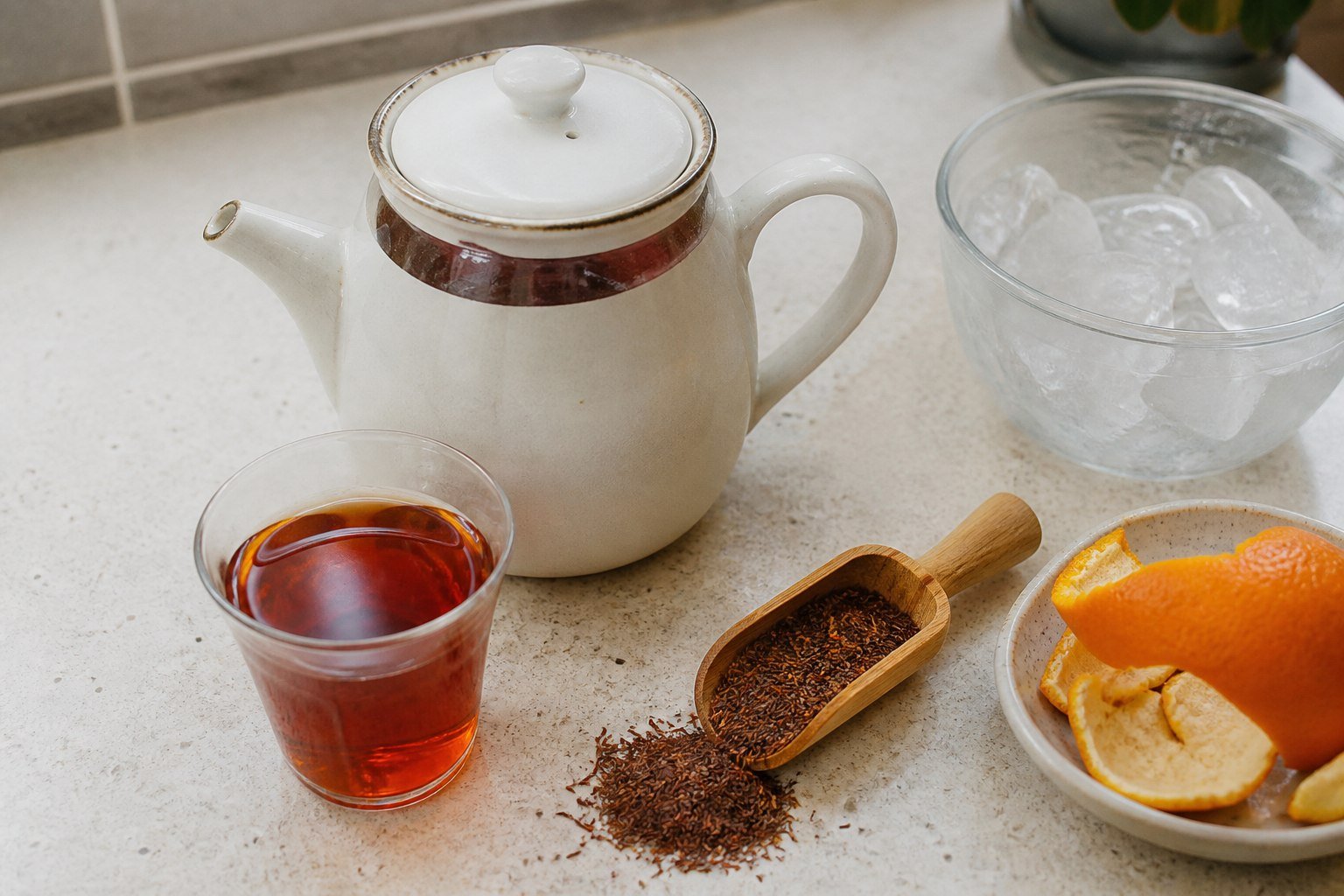 Rooibos tea steeping beside loose leaves, orange peel, and ice ready for cooling.