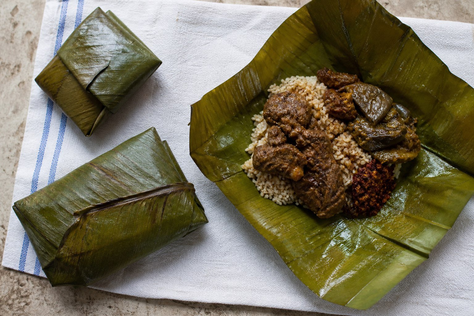Sri Lankan lamprais parcels beside an open banana leaf filled with rice, curry, eggplant, and sambol.