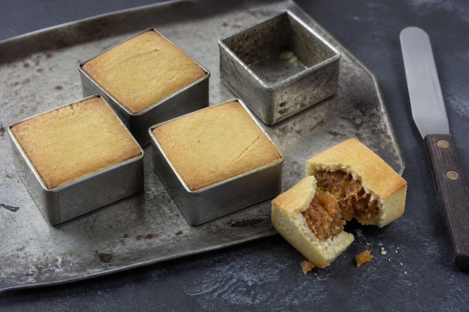 Taiwanese pineapple cakes cooling in square metal moulds beside a broken cake showing pineapple filling.
