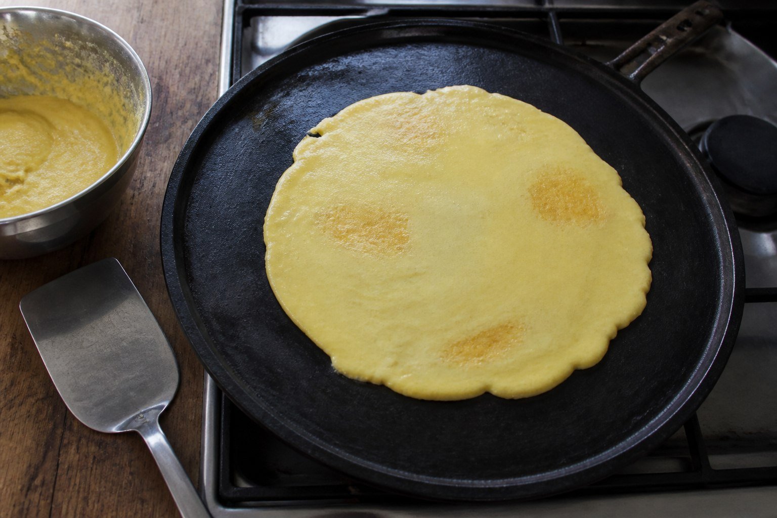 A smooth fresh-corn cachapa cooking on a cast-iron griddle beside a bowl of batter.