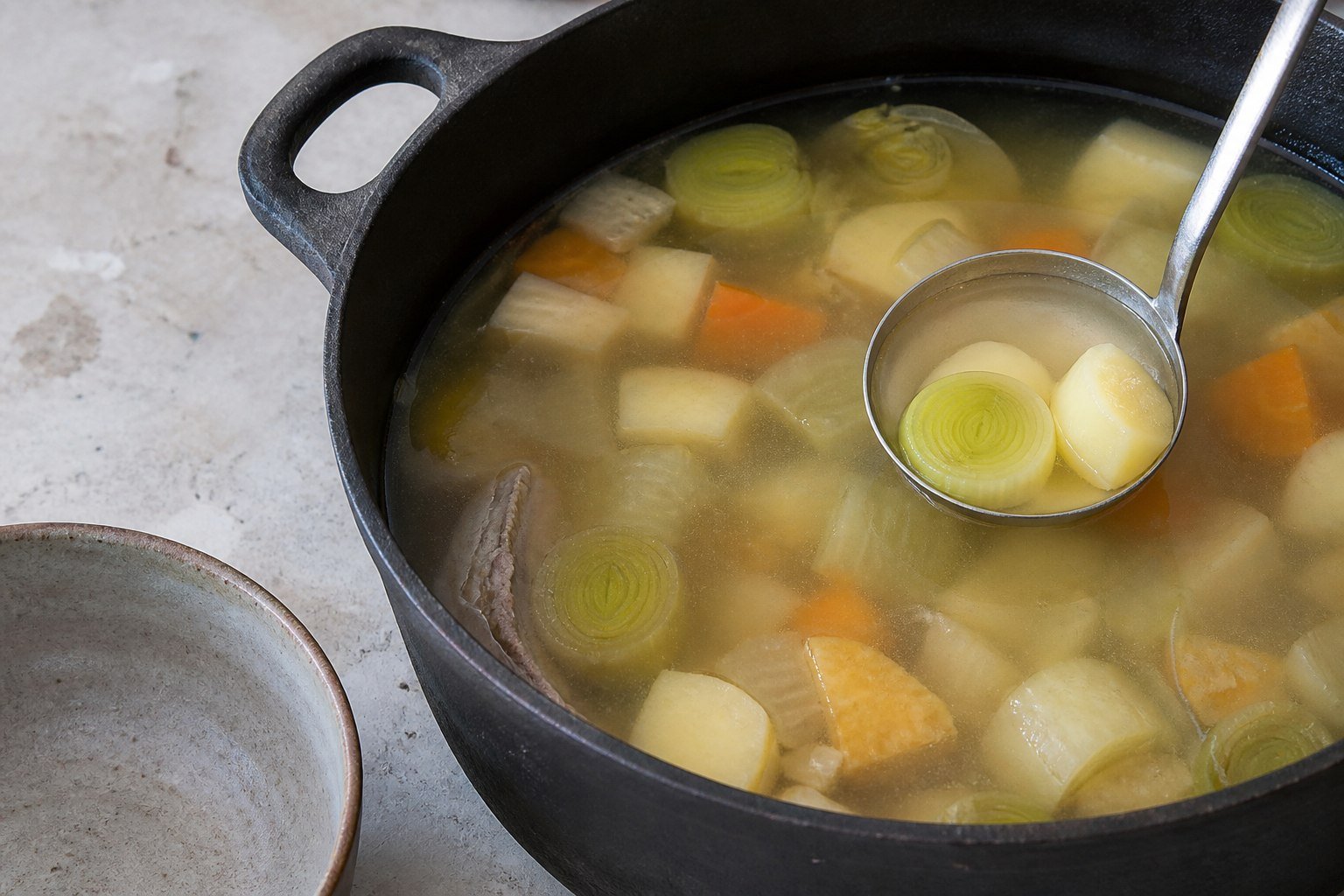 Welsh cawl being ladled from a cast-iron pot with leeks and potatoes visible in clear broth.