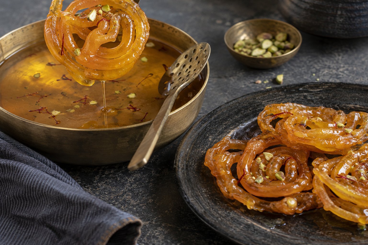 Indian jalebi being lifted from saffron syrup beside a slotted spoon and pistachios.