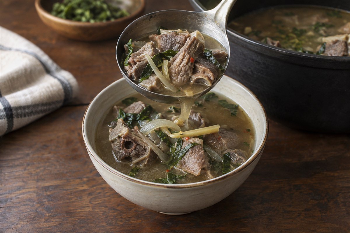Tender goat pieces and aromatic broth being ladled into a bowl of pepper soup.