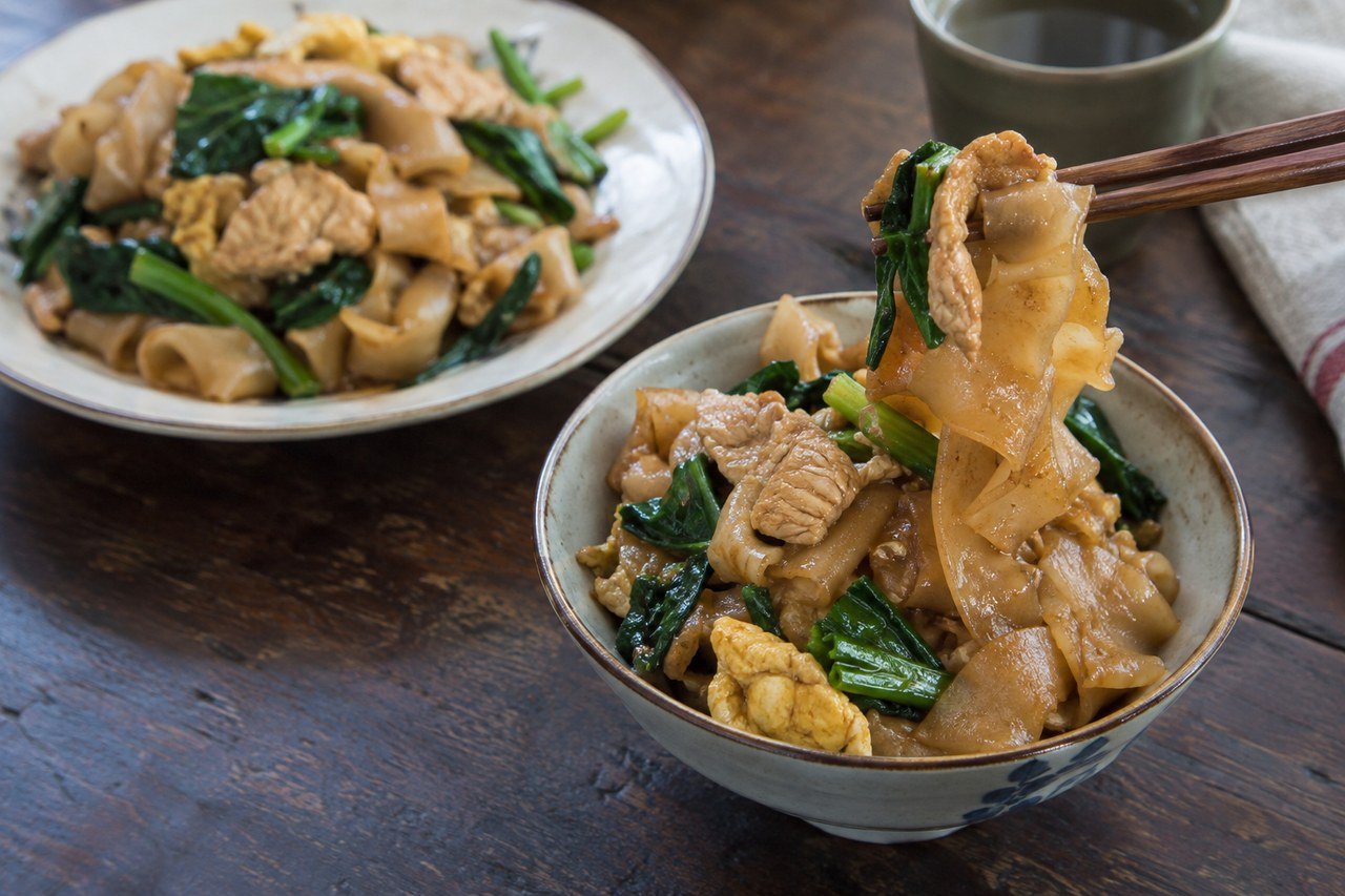 A serving of Thai pad see ew lifted with chopsticks beside a finished bowl of noodles.