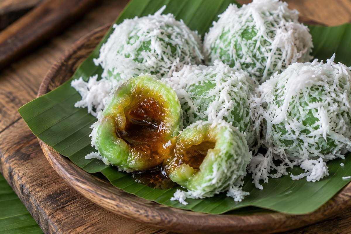 A close serving view of an Indonesian klepon being split open to show the melted palm sugar center, with coconut flakes and several whole rice cakes nearby.