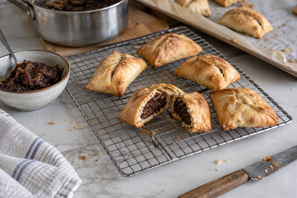 Maltese imqaret cooling on a wire rack beside date filling, a pastry knife, and unbaked pastries on a tray.