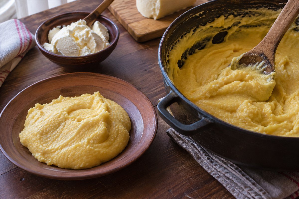 Montenegrin kačamak being served from a heavy pot onto an earthenware plate, with a wooden spoon, kajmak, and cheese nearby.