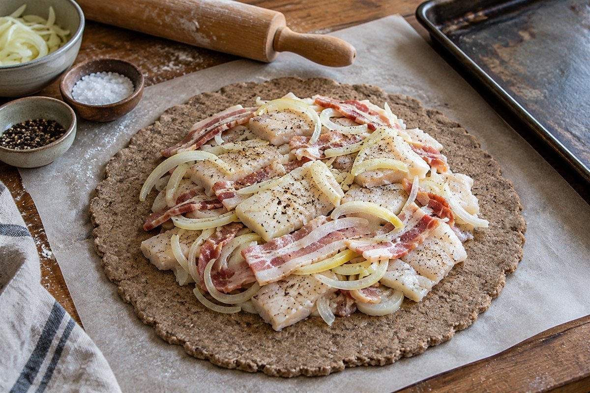 A Finnish kalakukko being sliced to reveal layers of white fish, pork, and onion inside the dark rye crust, with a linen napkin and knife nearby.