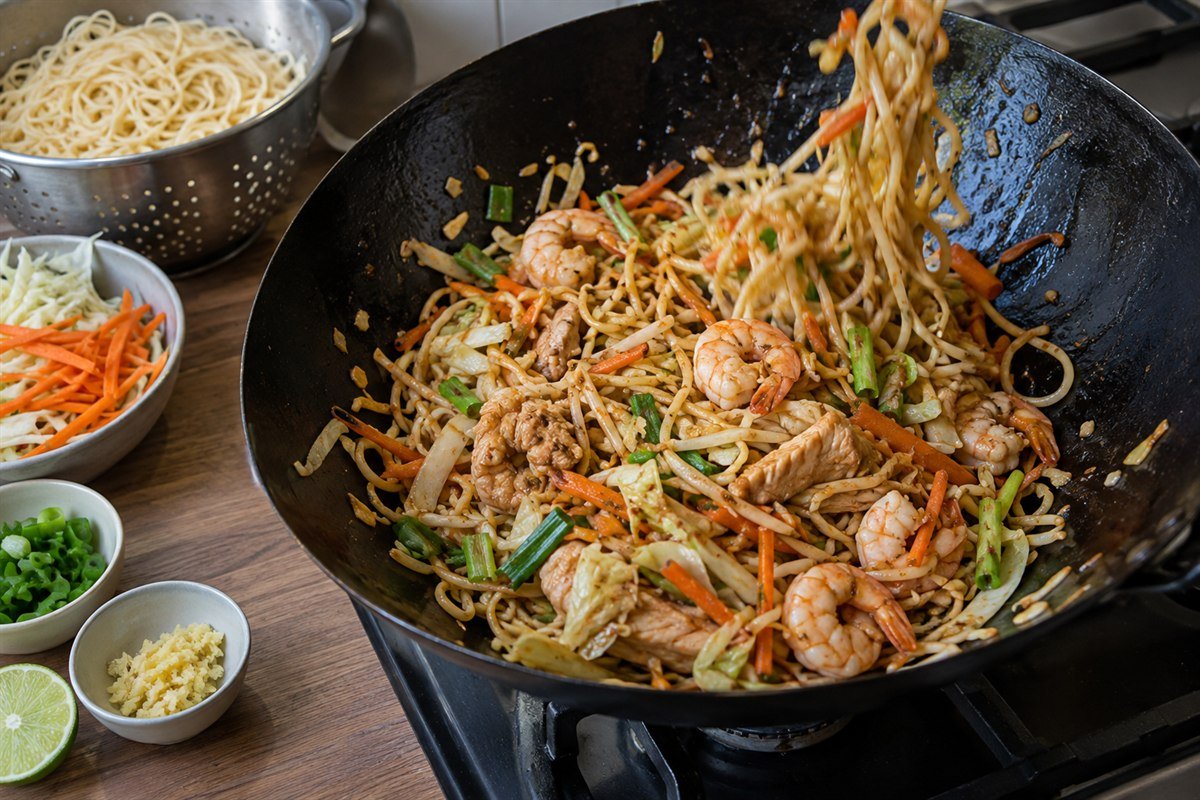 A wok of Mauritian mine frit being tossed with noodles, chicken, shrimp, ginger, and vegetables on a home kitchen work surface.