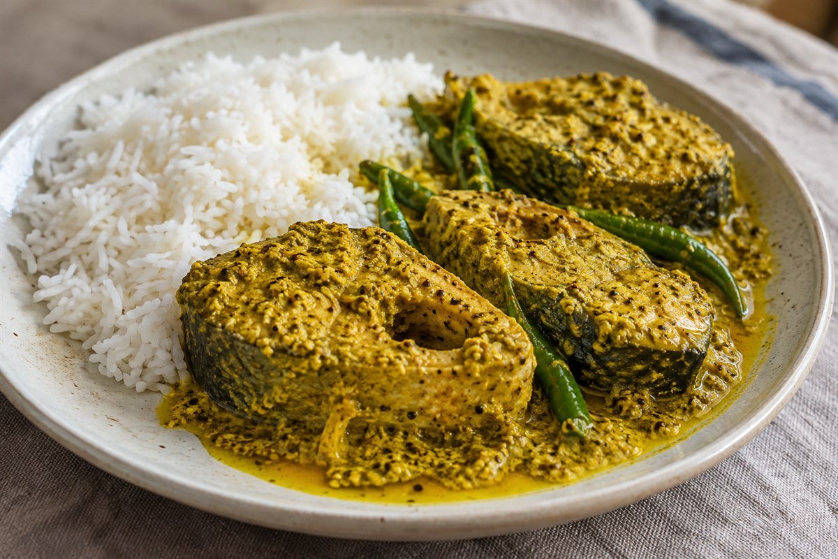 Bangladeshi shorshe ilish, thick pieces of hilsa fish in a golden mustard sauce with green chiles, mustard oil, and steamed rice on a simple plate in soft natural light.