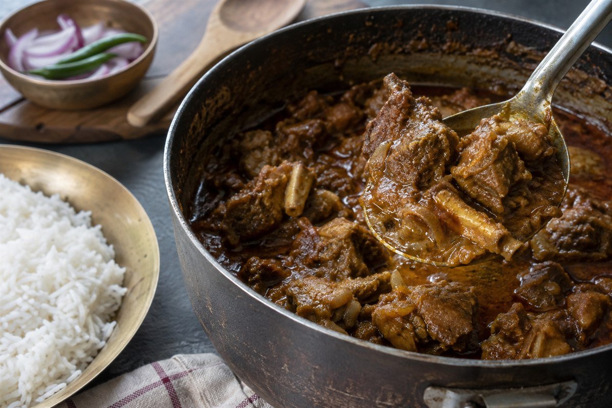 A spoon lifting tender goat from a pot of Bangladeshi kosha mangsho.