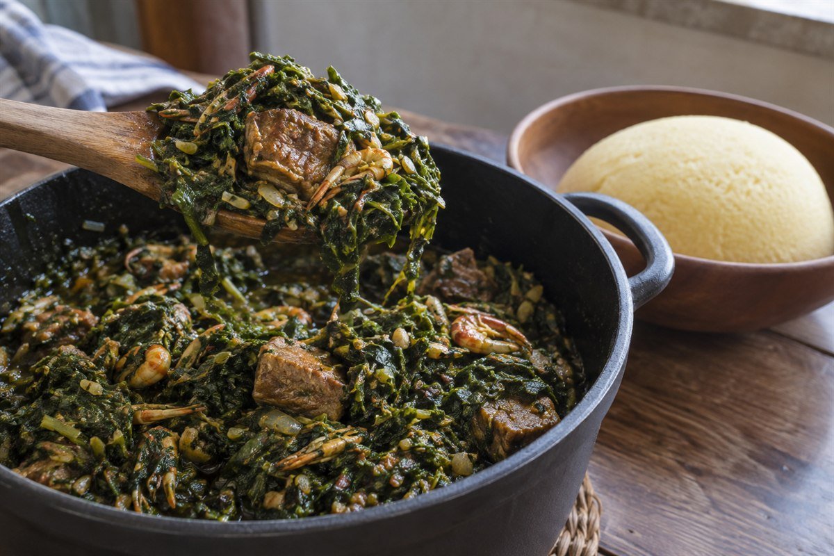 Cameroonian eru leafy greens stew being lifted from a pot beside soft garri.