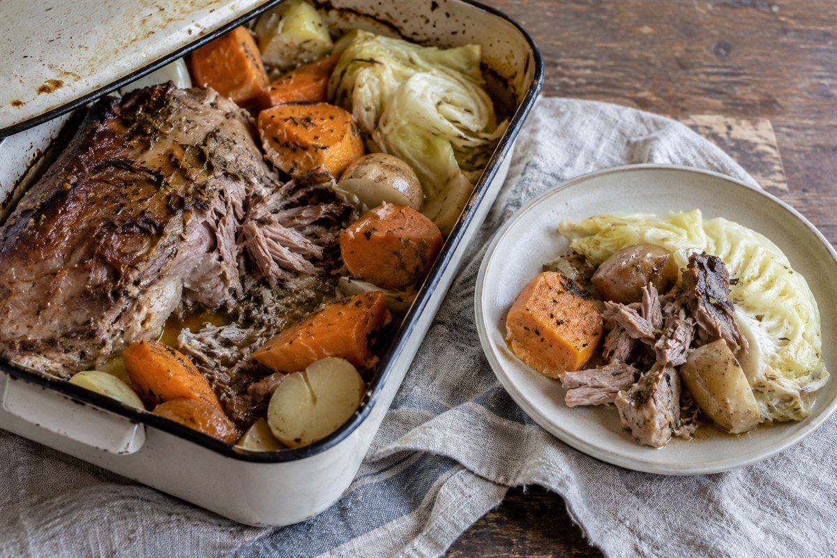 An oven pan and plate of New Zealand hangi-style pork, kūmara, potatoes, and cabbage ready to serve.